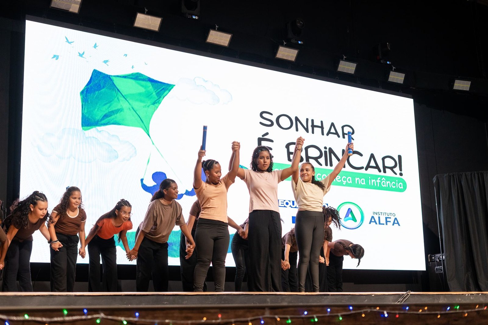 Adolescentes do Instituto Alfa em apresentação cultural no palco da campanha 'Sonhar é Brincar', com a logo do Instituto Alfa ao fundo.